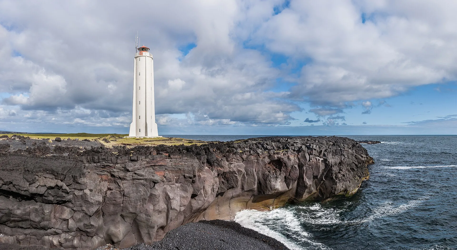 Der weiße Leuchtturm von Malarrif auf schwarzen Lavaklippen über dem Meer, Wolkenhimmel