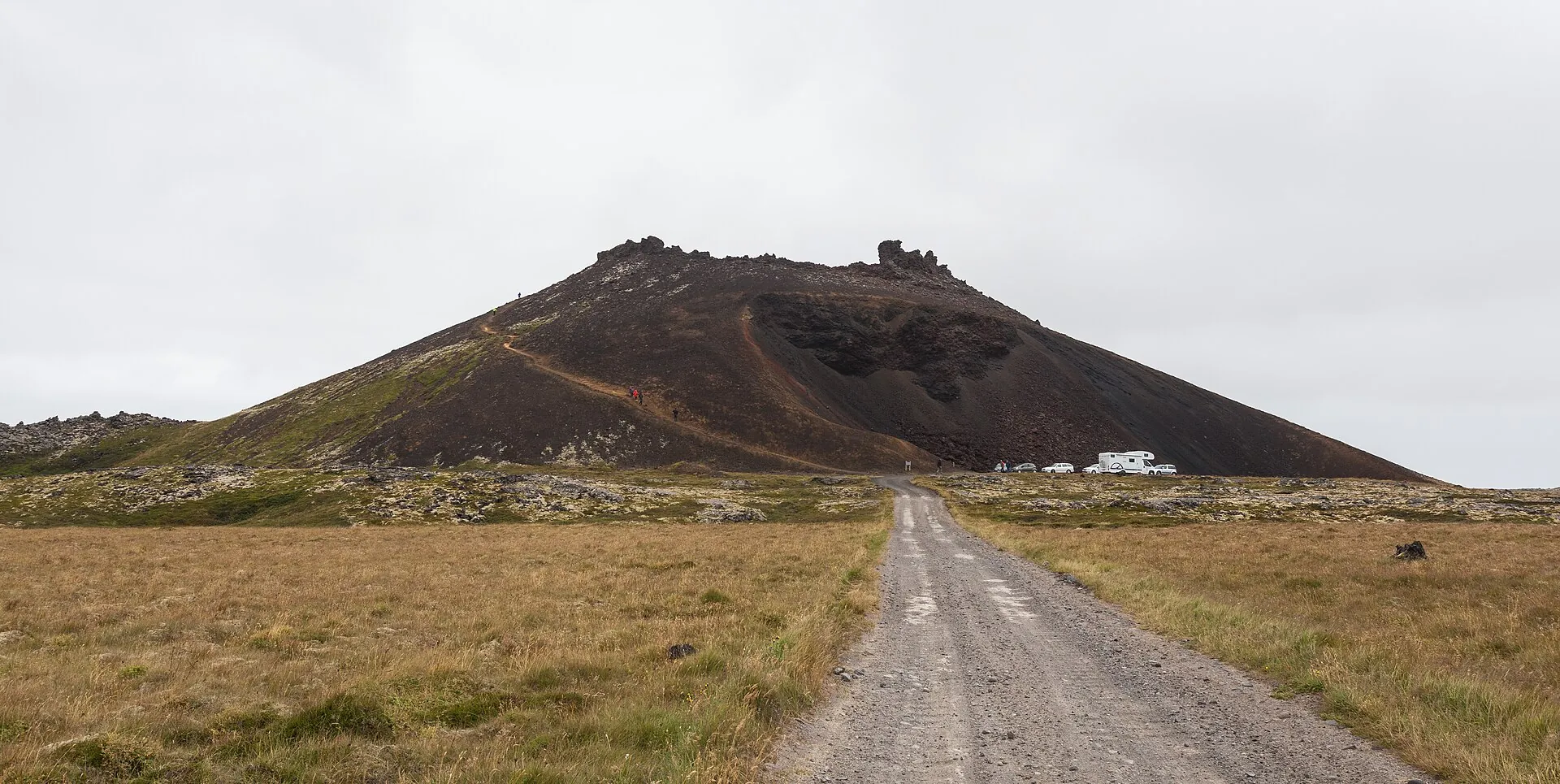 Der rotbraune Schlackenkegel Saxhóll mit der Stahltreppe zum Kraterrand, davor eine Schotterstraße durch Lavafeld