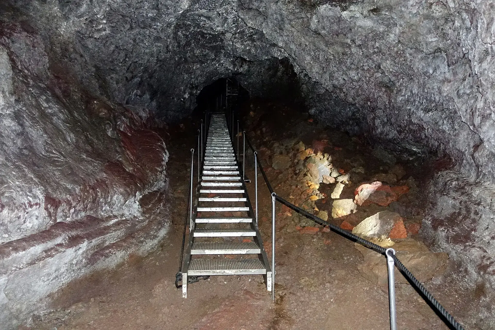 Die Stahltreppe hinab in die Lavahöhle Vatnshellir, rötliches Lavagestein, ein Geländer führt in die Dunkelheit