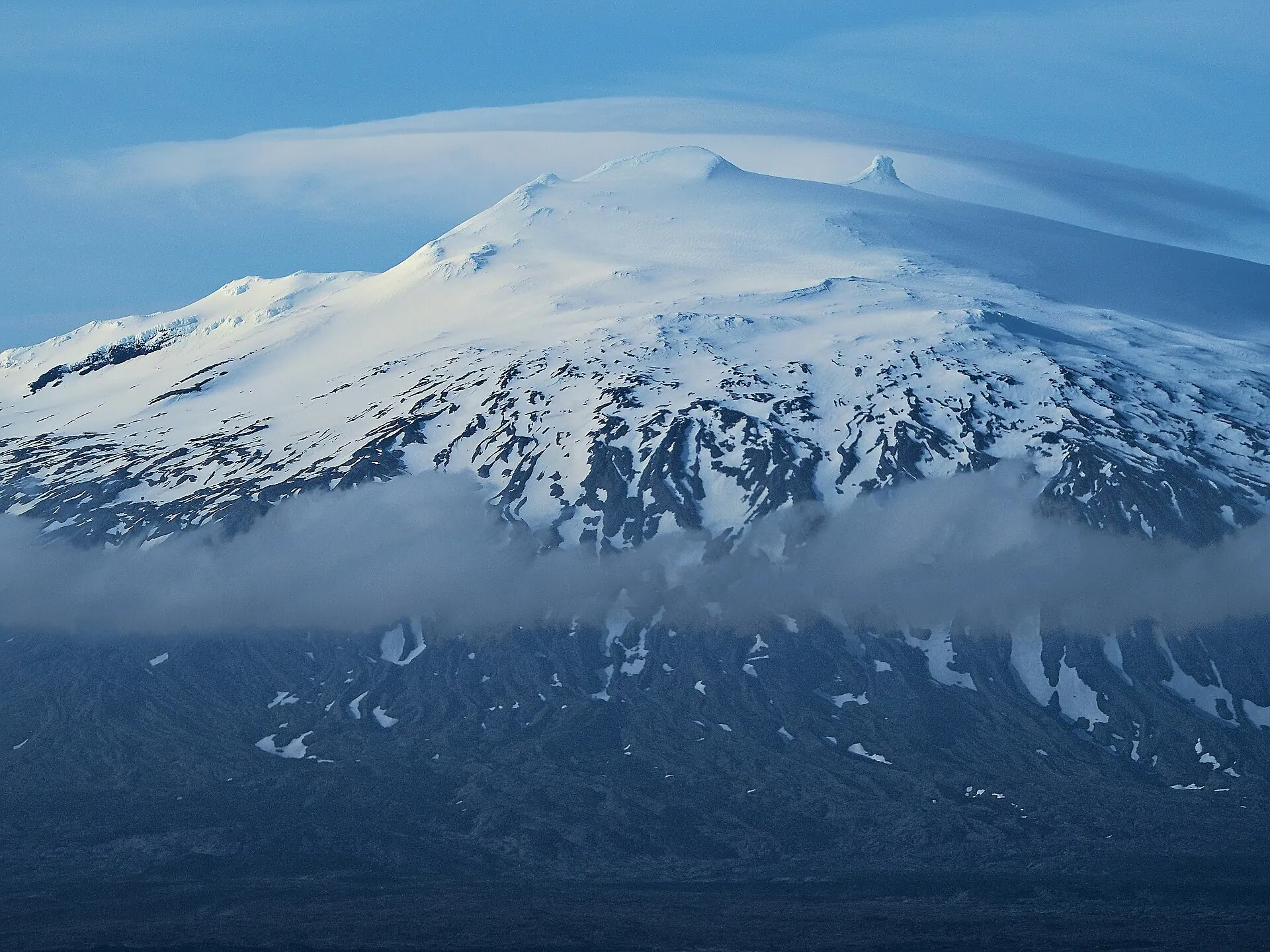 Der Snæfellsjökull mit einer Wolkenschicht am Hang und Wolken über dem Gipfel, blaugraues Abendlicht