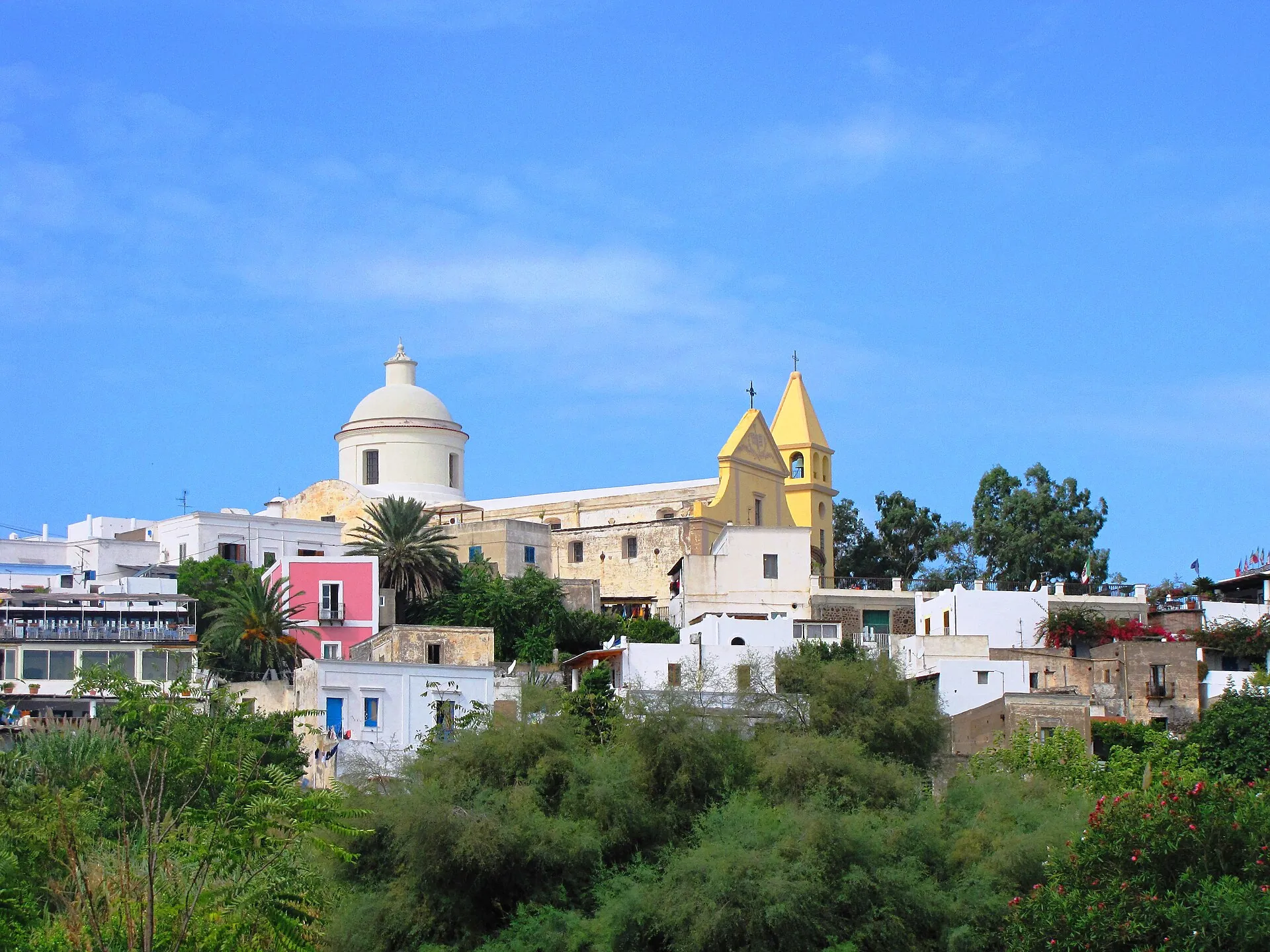 The white church of San Vincenzo with its yellow façade above the houses of Stromboli, palms and blue sky in front
