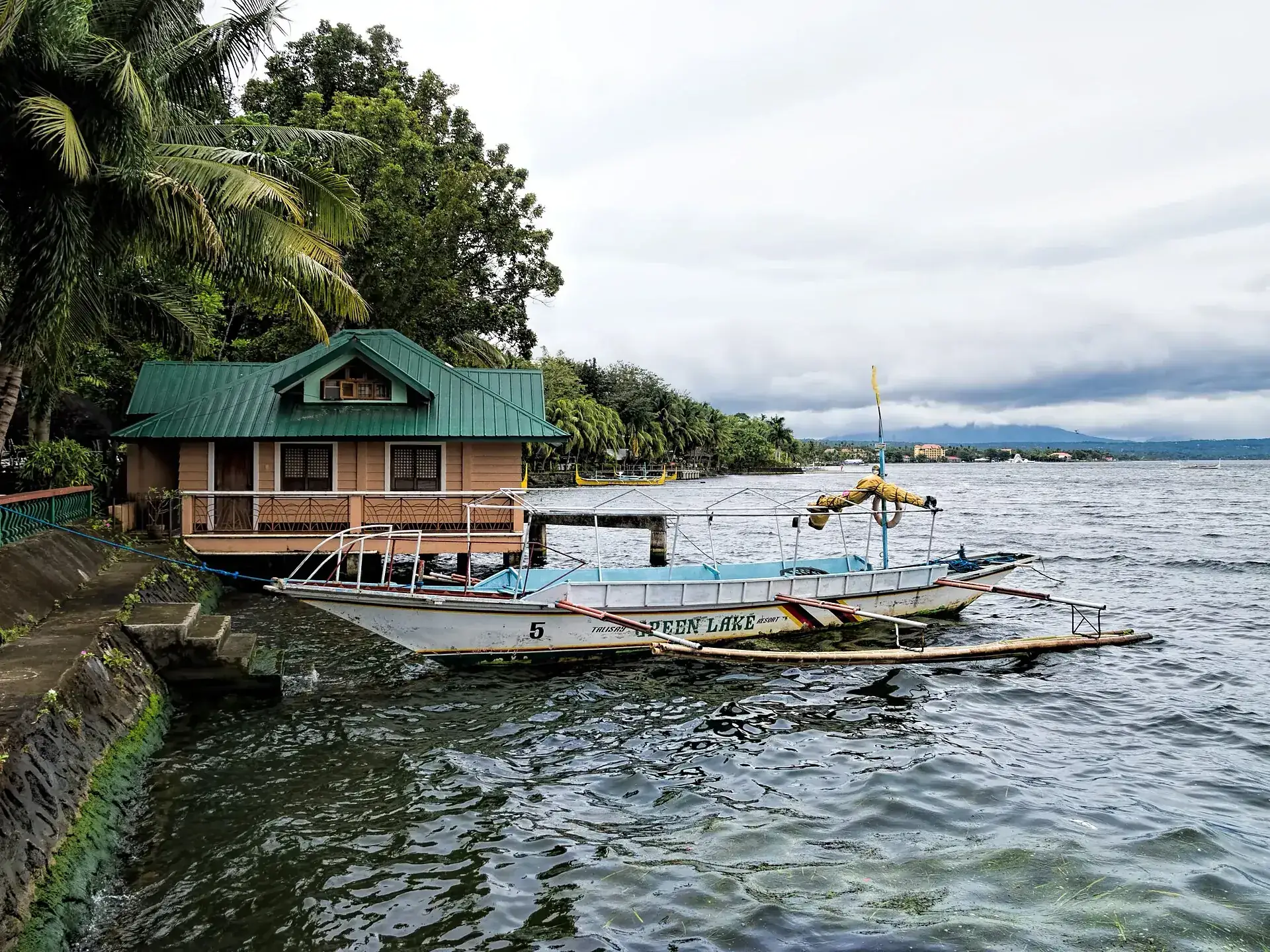Eine weiße Auslegerbangka mit blauem Rumpf liegt am Steg eines Resorts in Talisay am Taalsee, das Wasser leicht bewegt