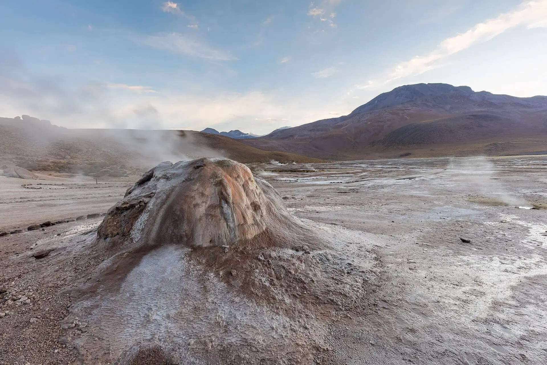 A reddish-brown sinter cone in the El Tatio geyser field, steam rising from its top, bare mountains behind