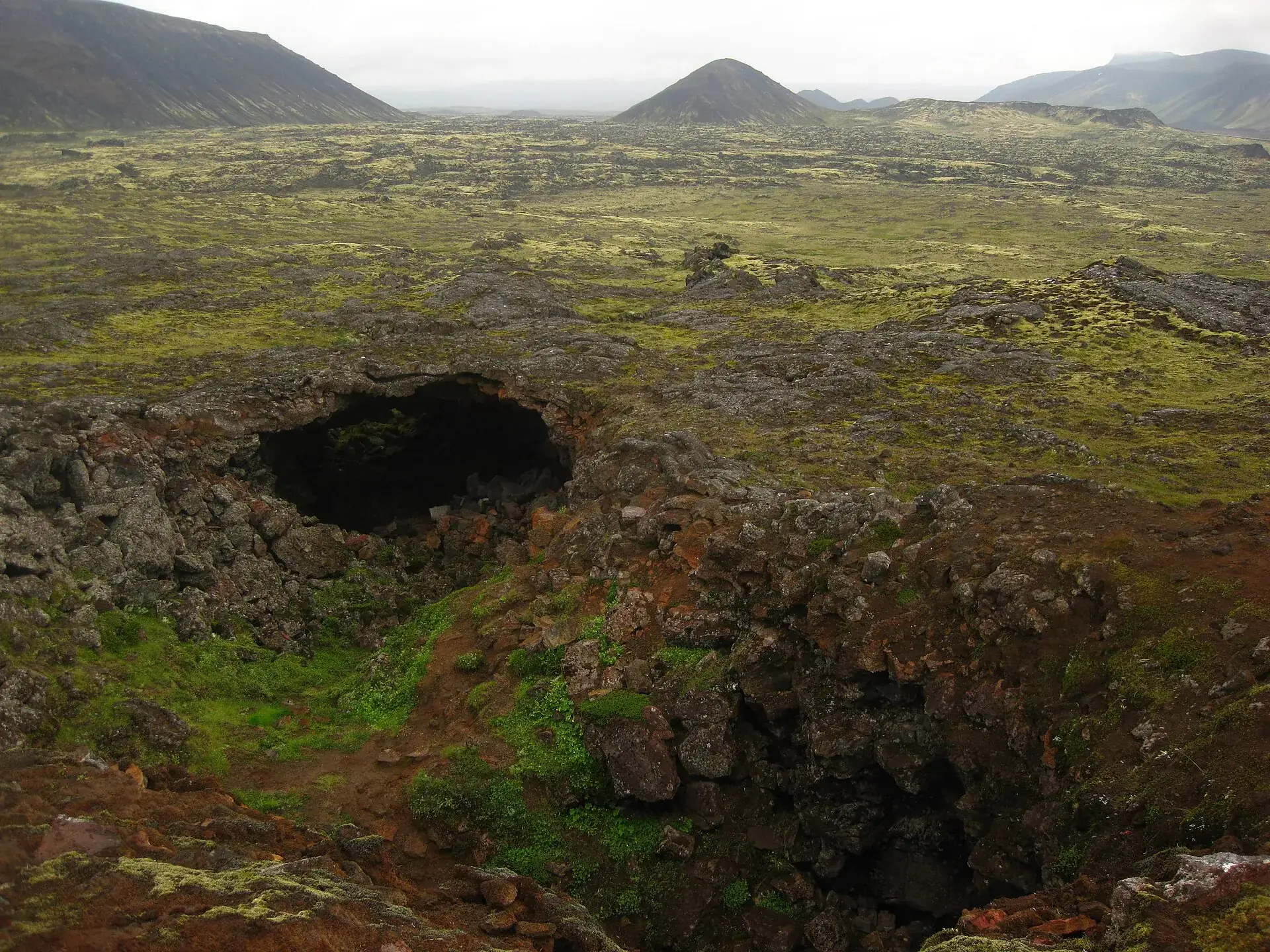 Moosbewachsenes Lavafeld in den Bláfjöll mit einer dunklen Senke im Vordergrund, dahinter Schlackenkegel unter Wolken