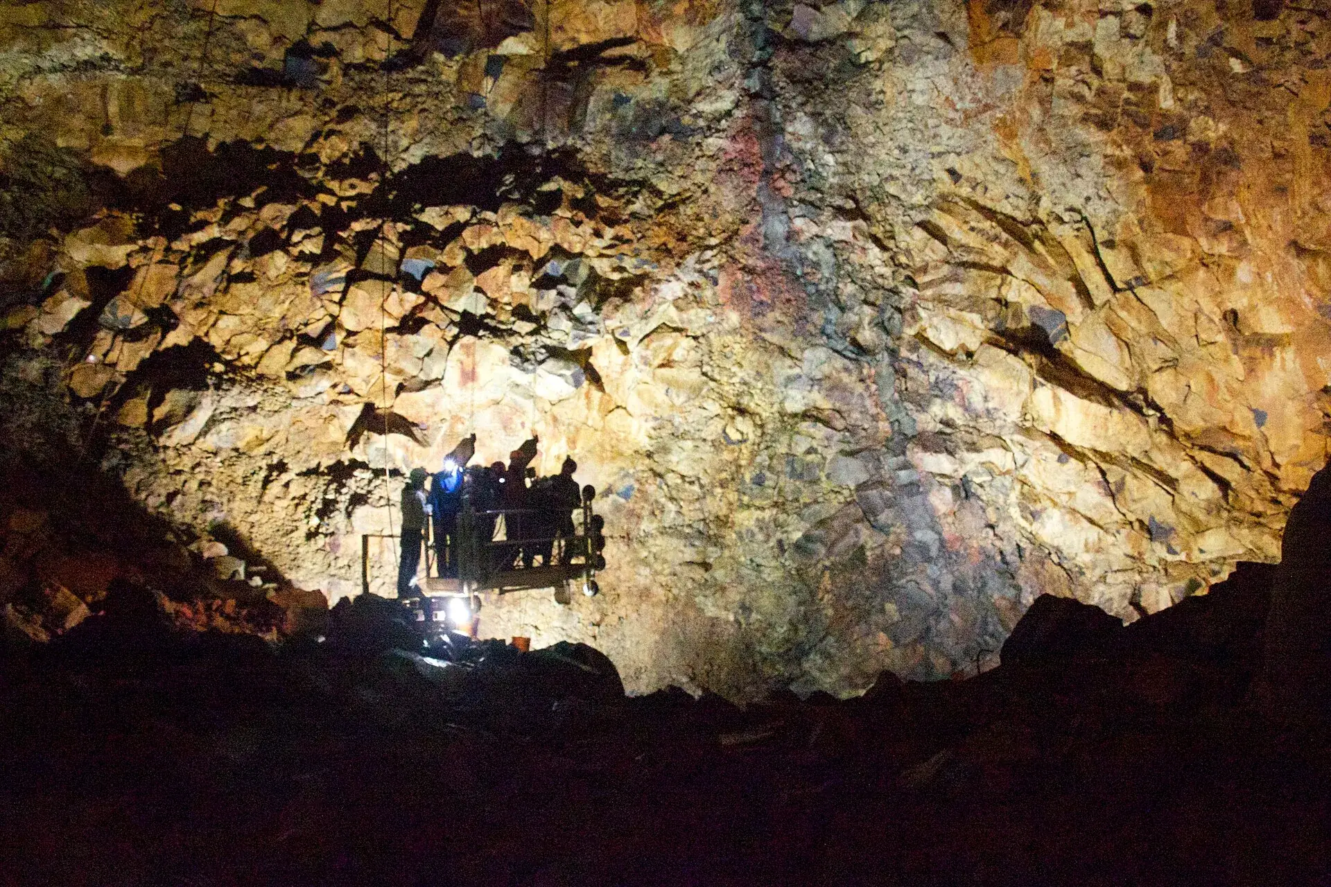 In der Magmakammer des Þríhnúkagígur: der offene Korblift mit Besuchern hängt vor einer bunt gefärbten Felswand, unten Scheinwerfer