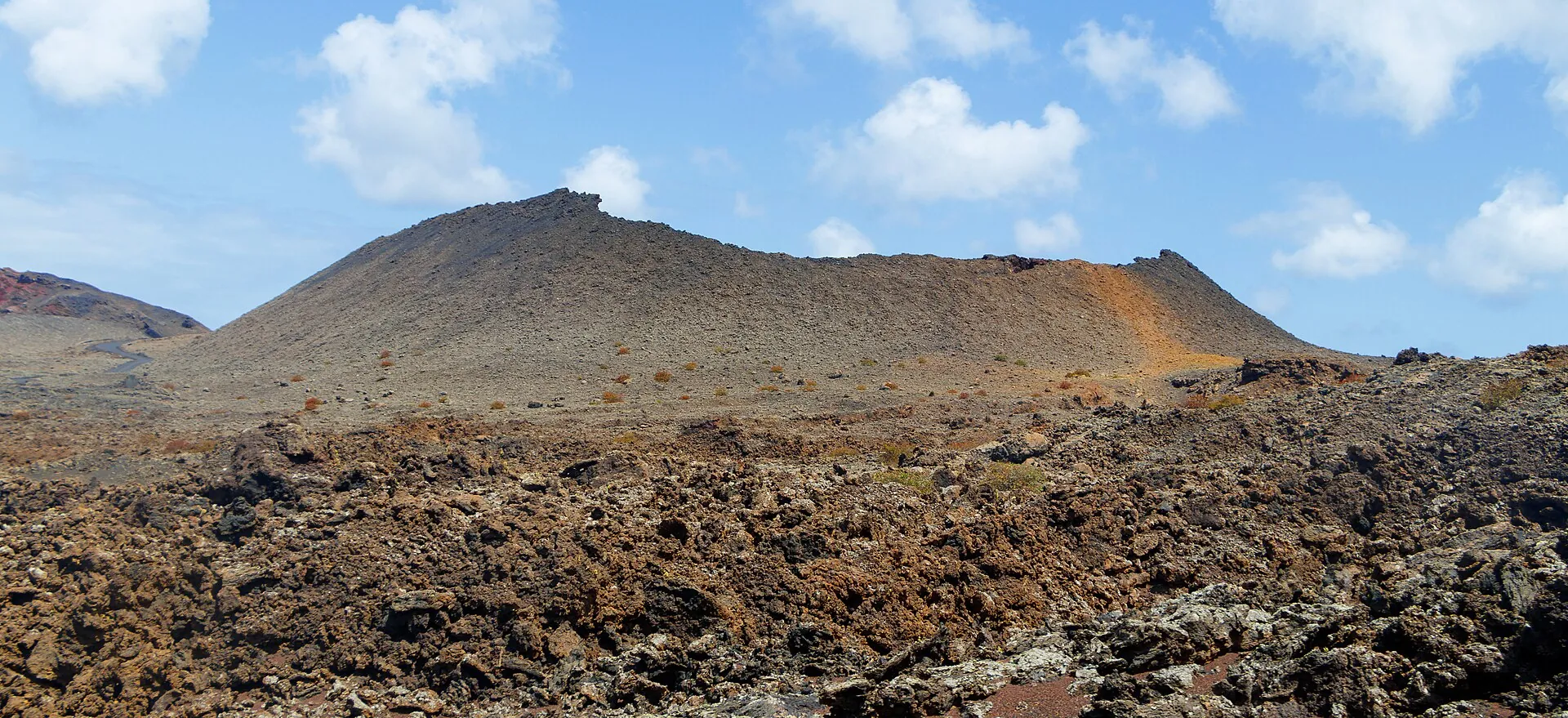 Wide lava plain in Timanfaya National Park, two flat volcanic cones behind under a blue sky