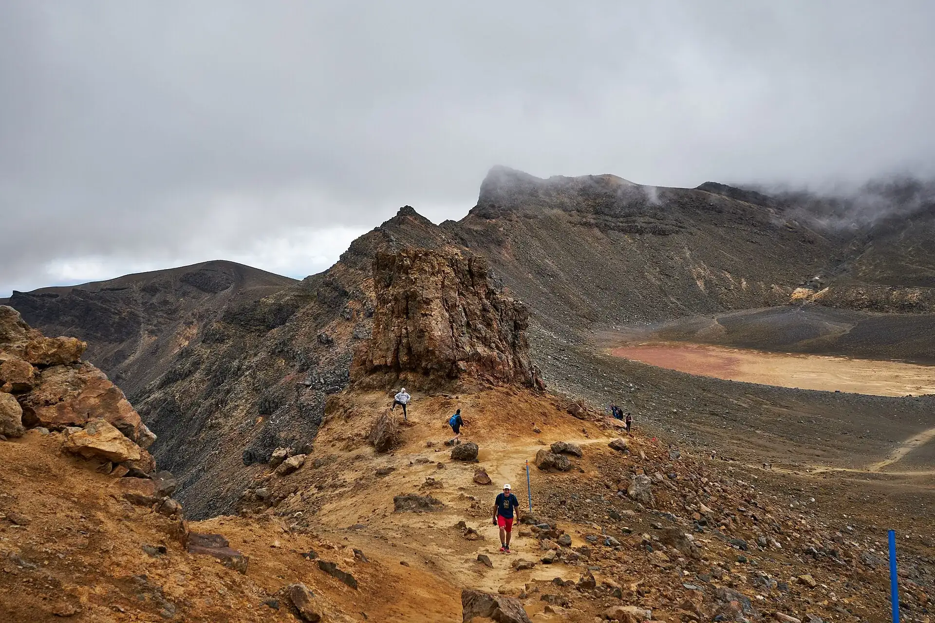 Walkers on the ridge at the Red Crater of the Tongariro Alpine Crossing: a narrow path over ocher rock, with the Central Crater behind under low cloud