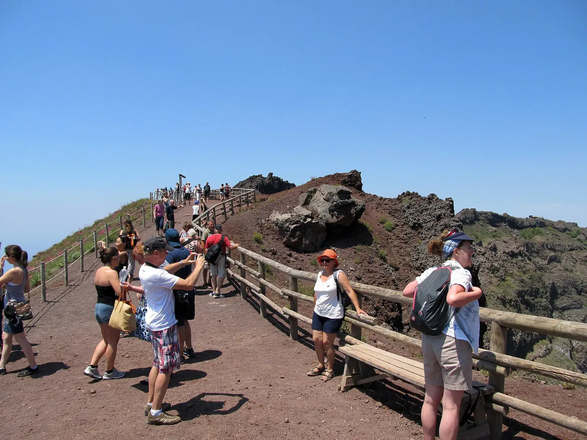 The path along the crater rim of Mount Vesuvius: reddish lava gravel, a wooden railing, visitors walking uphill