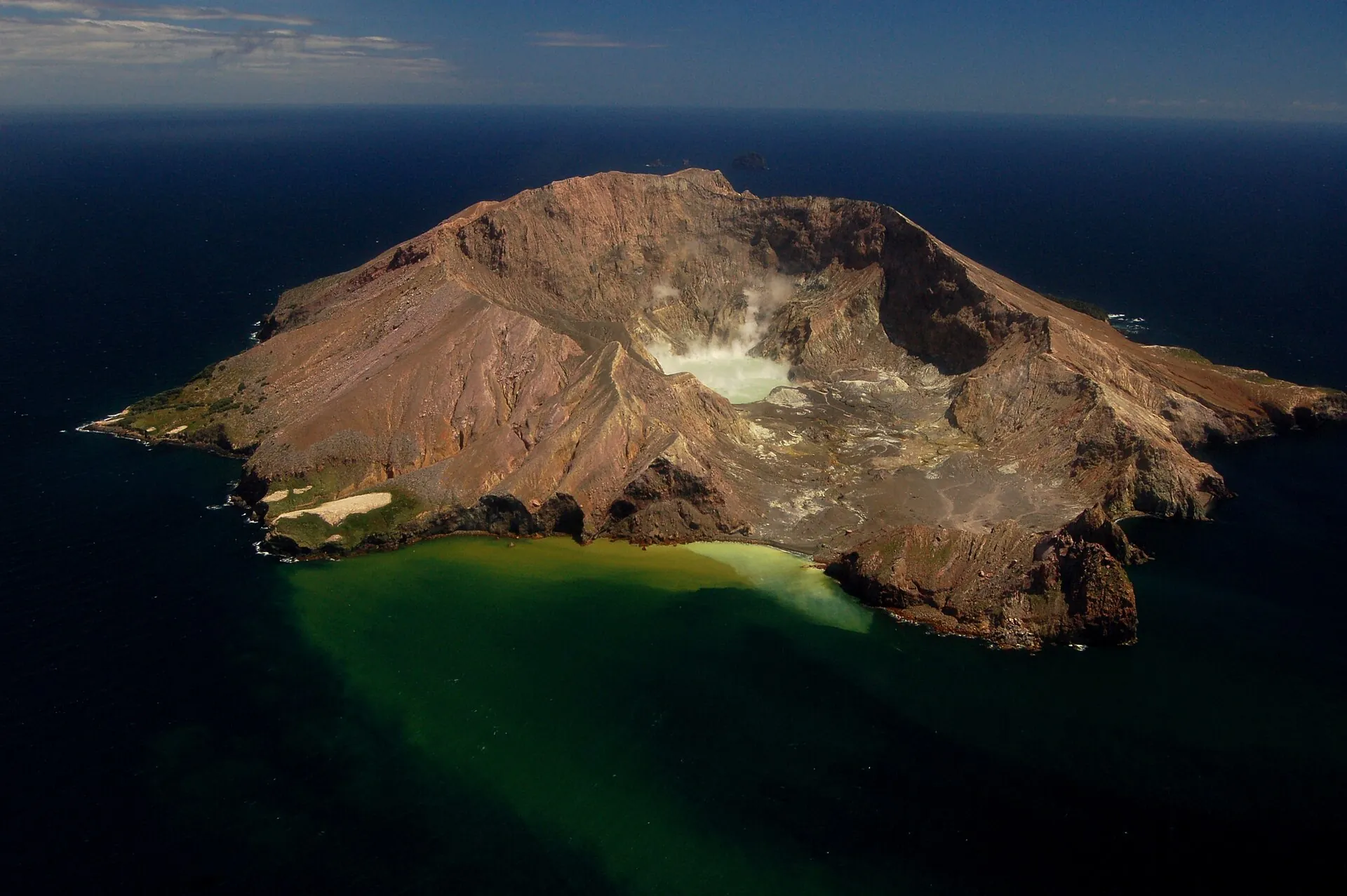 Whakaari / White Island from the air: the brown volcanic island in the sea, steam standing over the crater, the water in front stained yellow-green