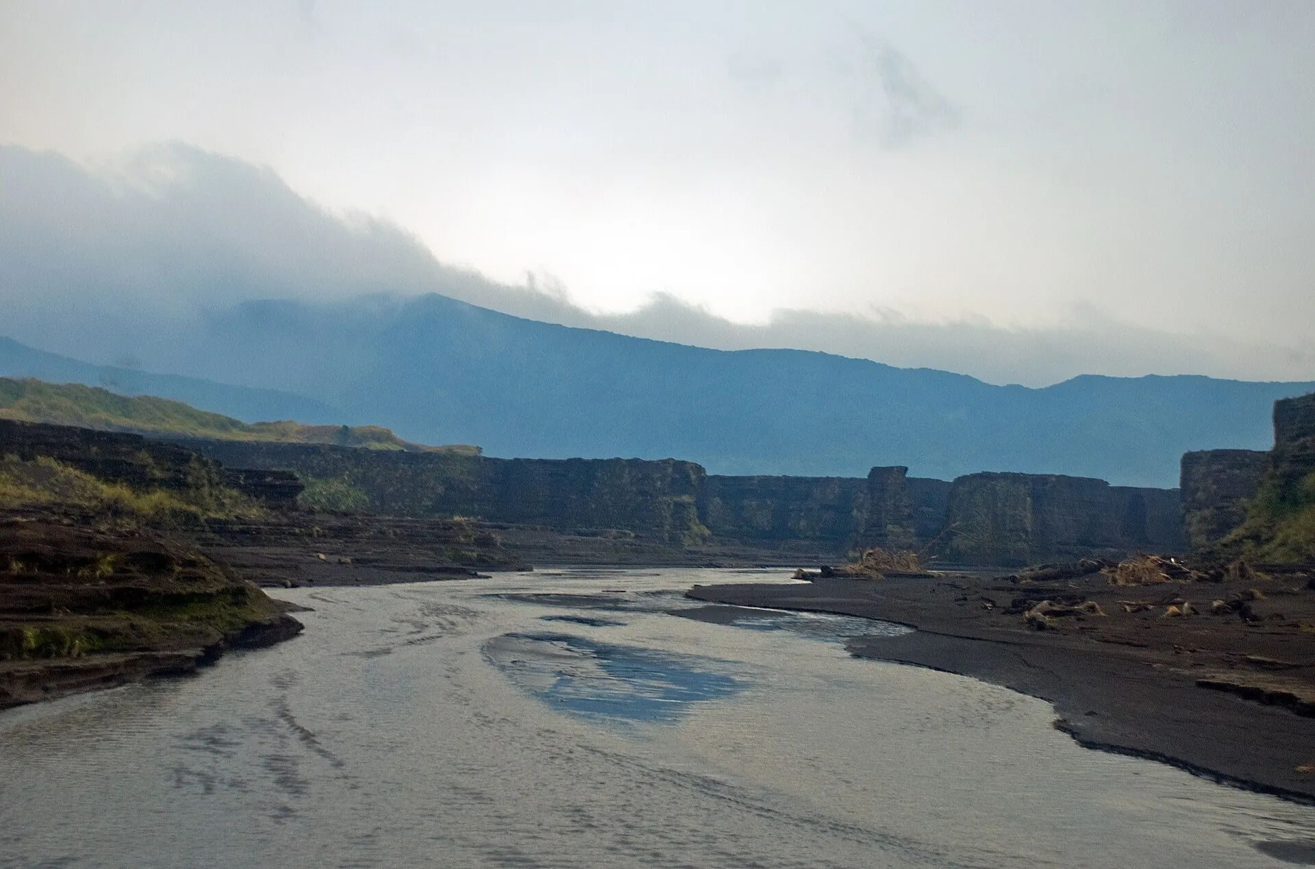 Ein flacher Wasserlauf zieht sich über die graue Ascheebene am Fuß des Mount Yasur, im Hintergrund bewaldete Hänge