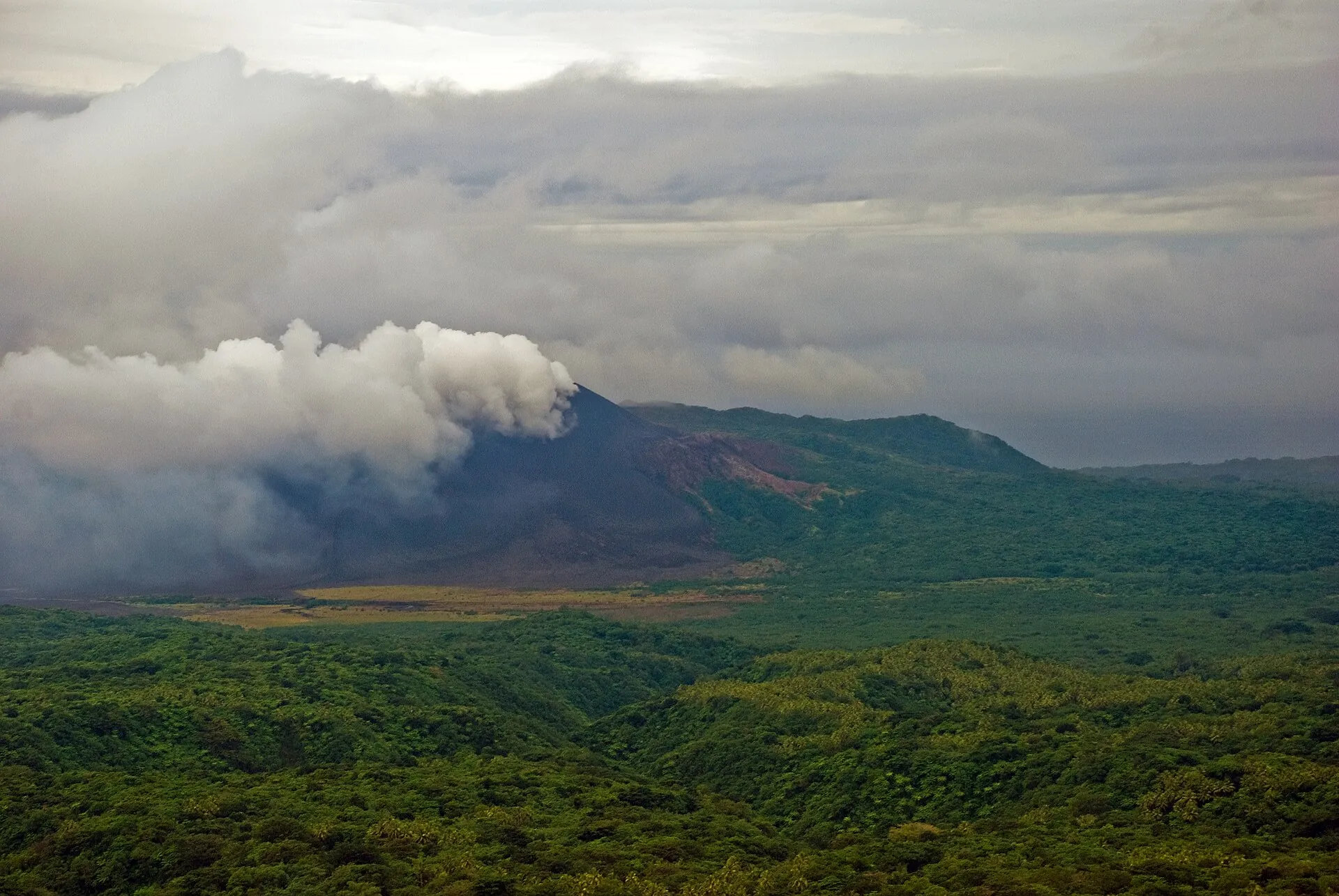 Der Kegel des Mount Yasur aus der Ferne über der Ascheebene von Tanna, darüber eine breite Dampffahne