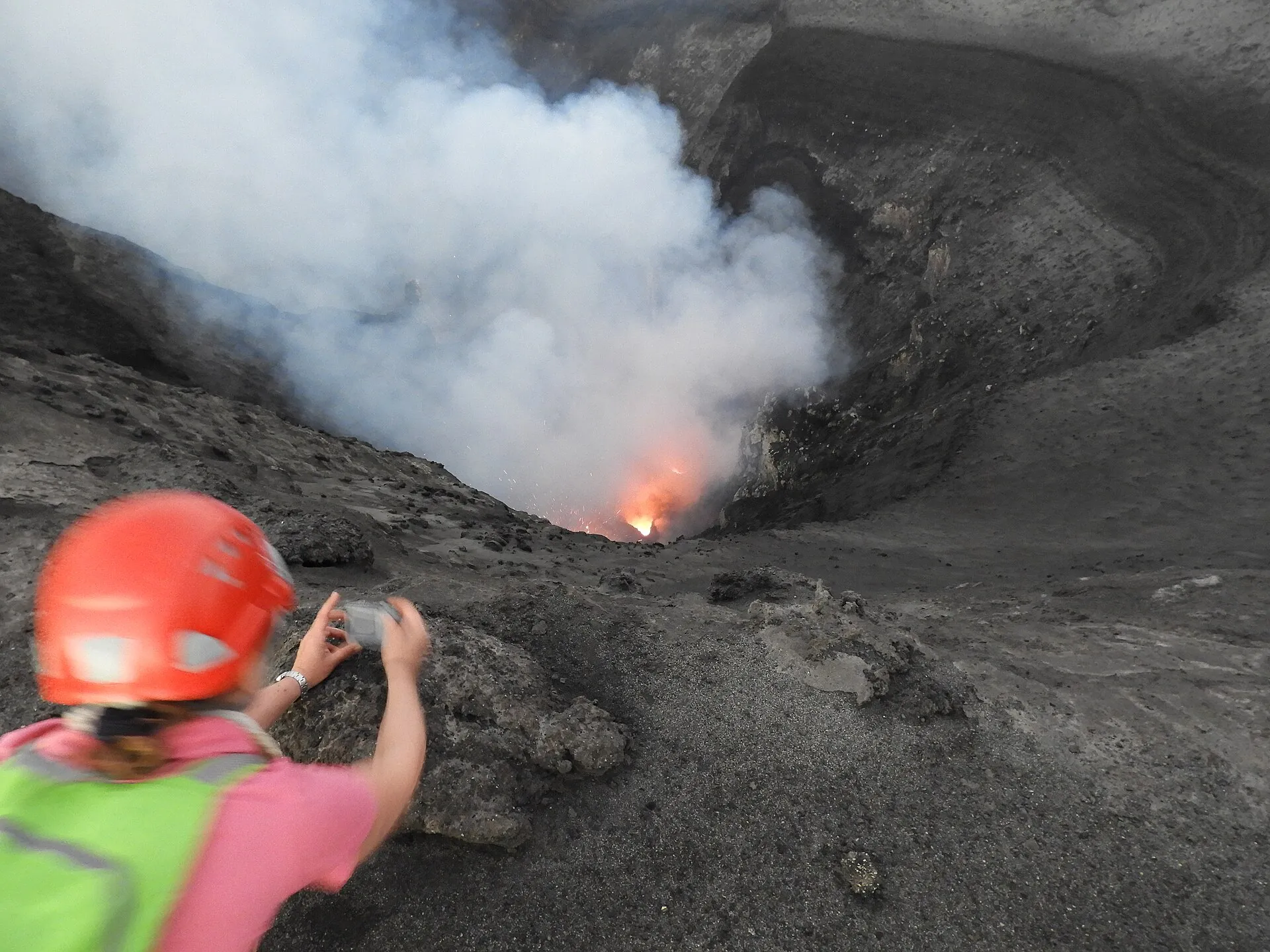Ein Besucher mit Schutzhelm steht in der Dämmerung am Kraterrand des Mount Yasur, hinter ihm steigt Rauch auf